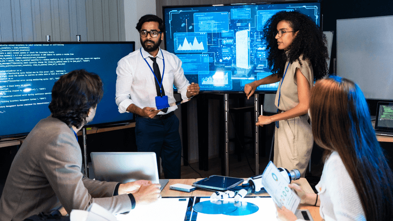 A diverse team collaborates around a table with screens displaying data and visuals.