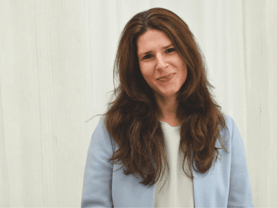 Woman with long brown hair in a light blue blazer stands smiling against a white background