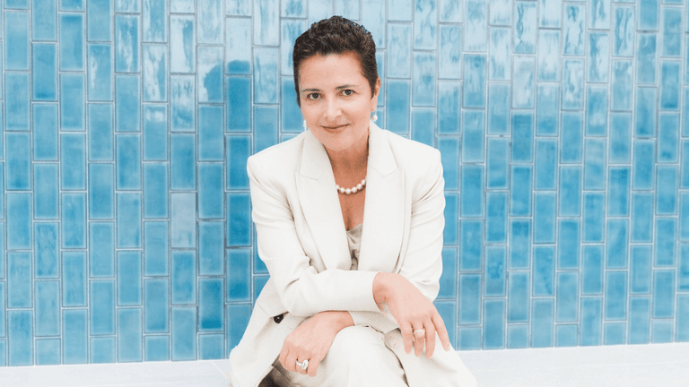 A woman in a cream suit sits against a blue tiled wall, smiling gently